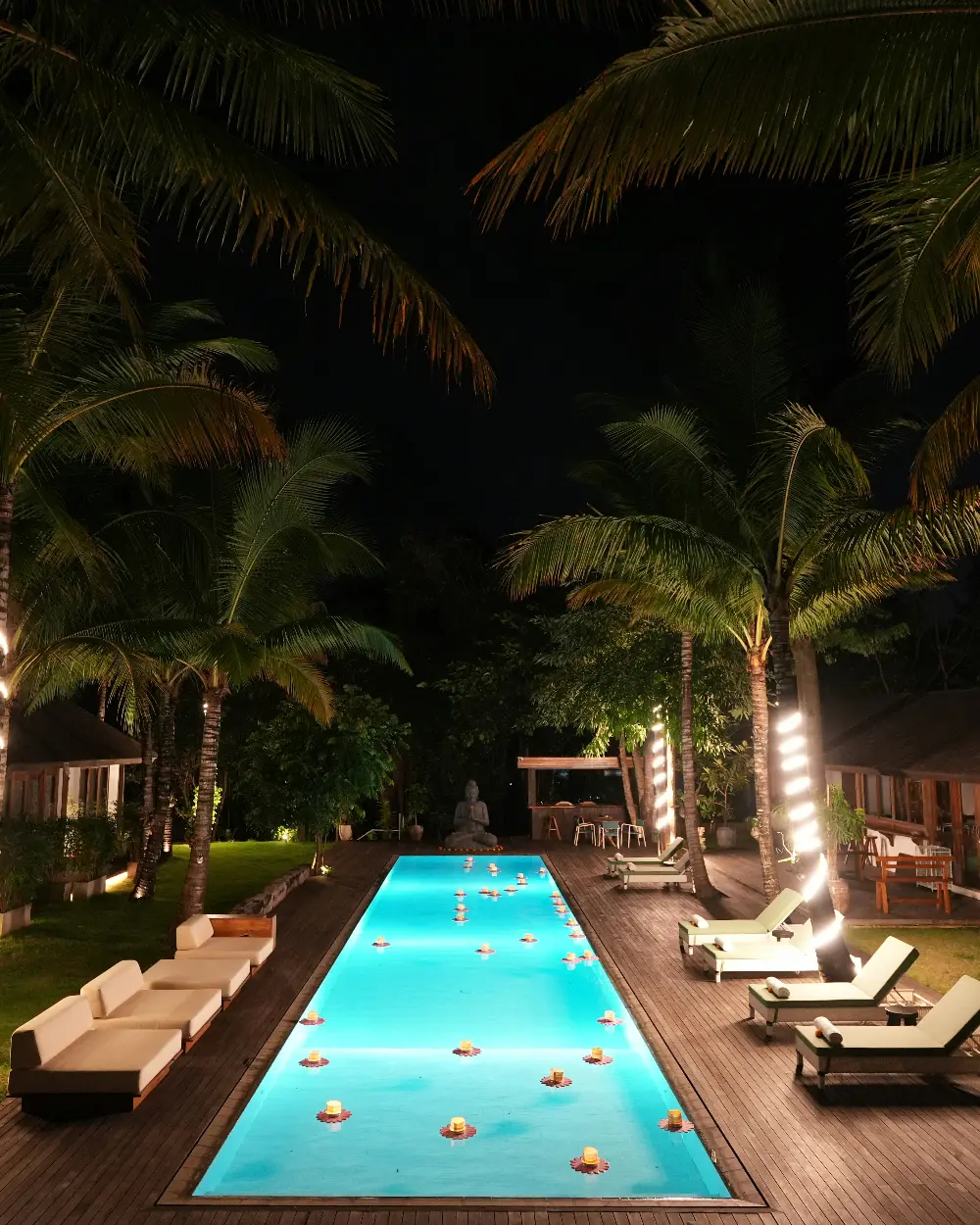 Piscina iluminada de noche, rodeada de palmeras y velas flotantes